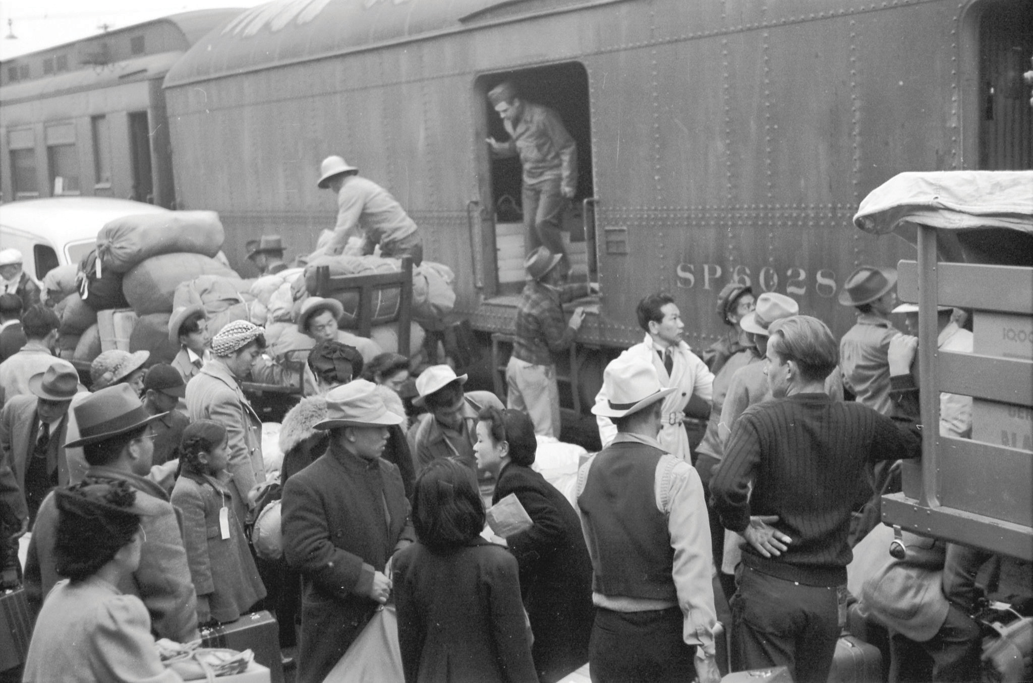 Black-and-white photo. Japanese Americans going to Manzanar gather around a baggage car at the old Santa Fe Station.