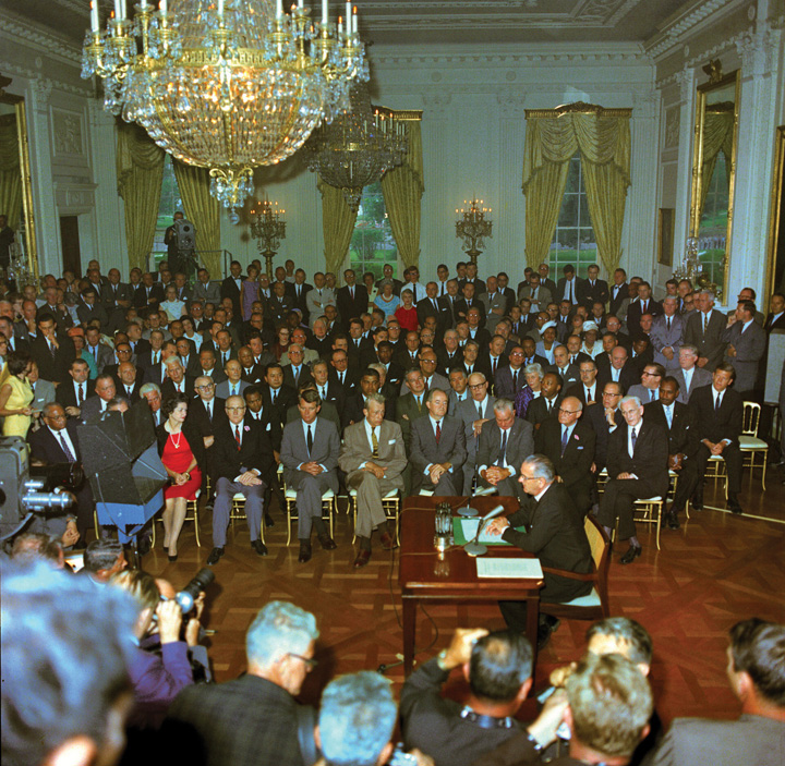 Photo of President Lyndon B. Johnson signing the Civil Rights Act in the East Room of the White House. People watching include Attorney General Robert Kennedy, Senator Hubert Humphrey, First Lady "Lady Bird" Johnson, Rev. Martin Luther King, Jr., F.B.I. Director J. Edgar Hoover, Speaker of the House John McCormack. Television cameras are broadcasting the ceremony.