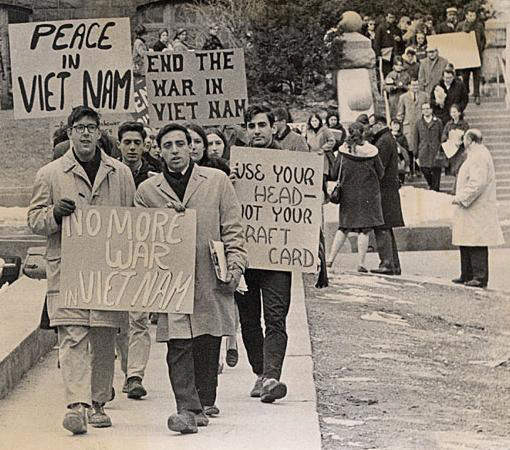 Black-and-white photo of students protesting the Vietnam War. Many are carrying signs and placards, with messages like "Peace in Vietnam" and "No more war in Vietnam."