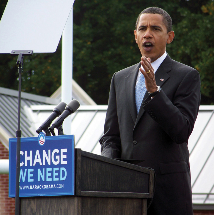 Photo of Barack Obama standing behind an outdoor podium, which holds a sign that reads, "Change we need."