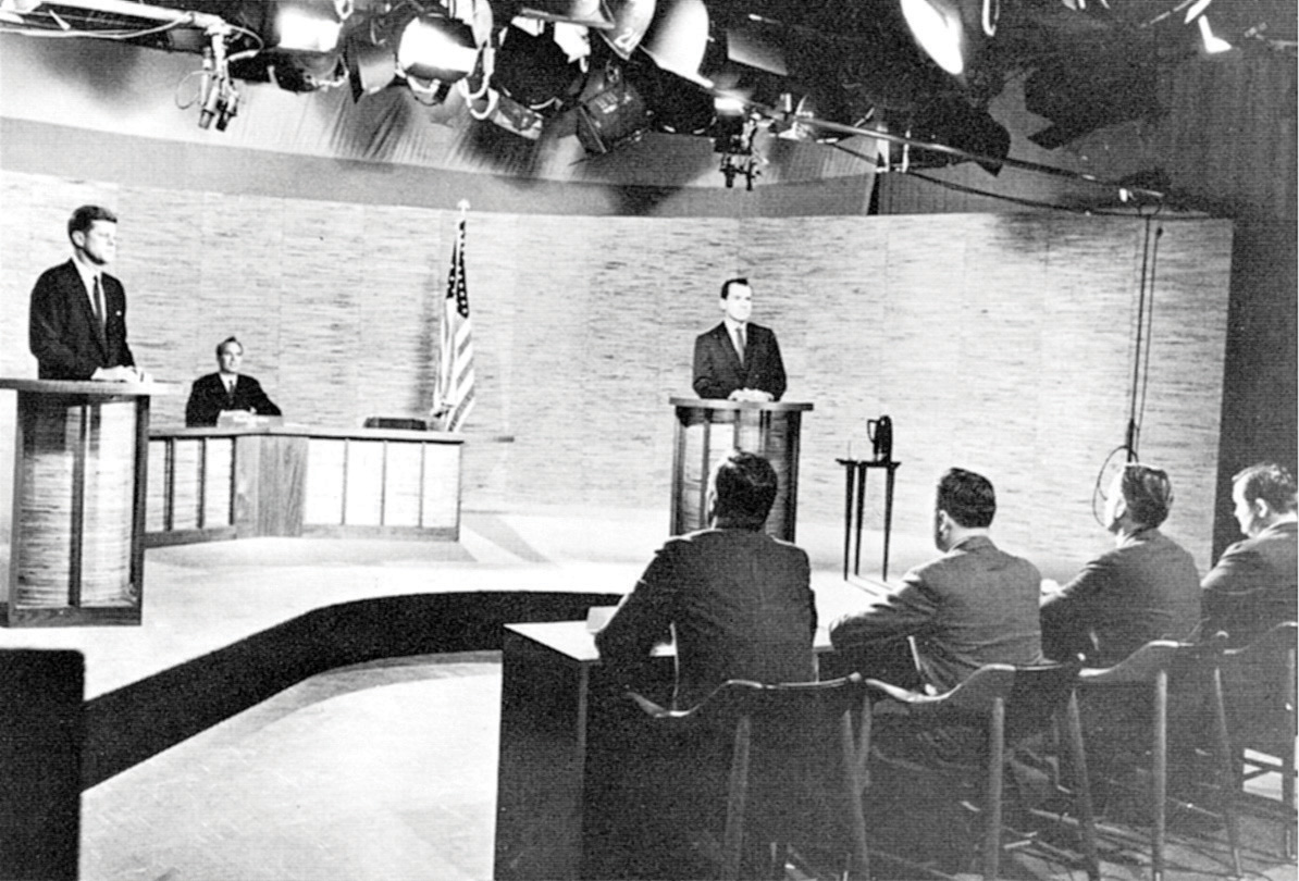 Black-and-white photo of John F. Kennedy onstage with Richard Nixon during a debate.