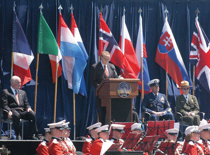 Photo of Bob Dole onstage behind podium. Stage is filled with national flags from around the world, and just below the stage, in front, are members of a military band.
