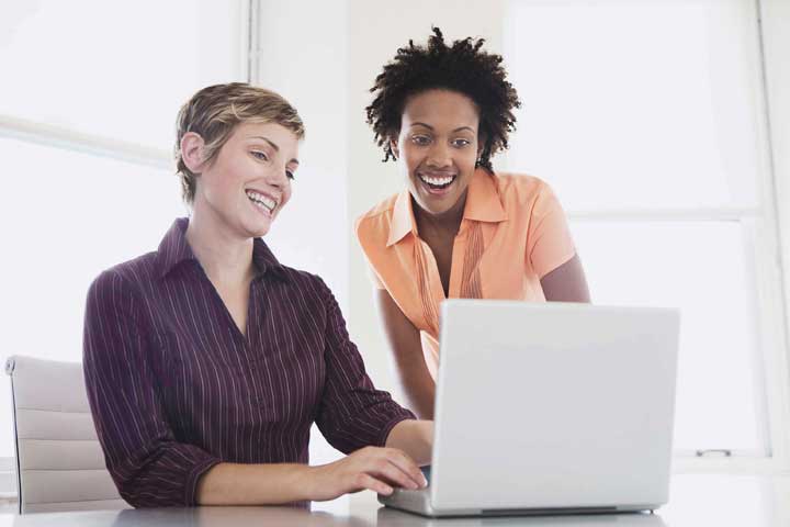 Person at a laptop talking happily with person looking on from the side